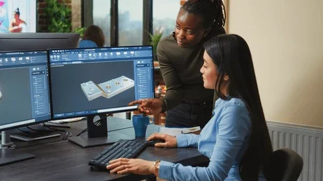 A woman at a desk using a computer displays 3D design software, while a colleague points at the screen in a modern office setting.