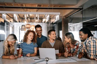A diverse group of six young professionals gathered around a laptop, collaborating and reviewing papers in a modern office setting.