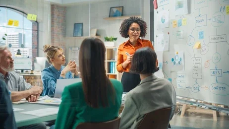 A woman stands by a whiteboard, presenting to a group of four colleagues seated at a conference table in a modern office setting.