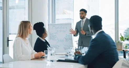 A diverse group of professionals sits around a table, attentively listening to a speaker presenting a marketing strategy on a whiteboard.